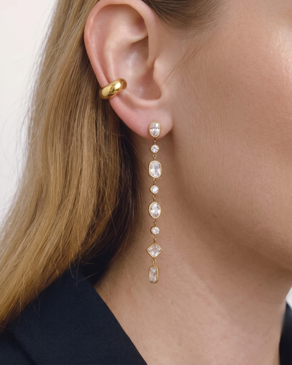 close-up photograph of a long gold dangle earring featuring varied-cut clear crystals (oval, round, and diamond shapes), and a thick gold ear cuff, both worn on a woman's ear.