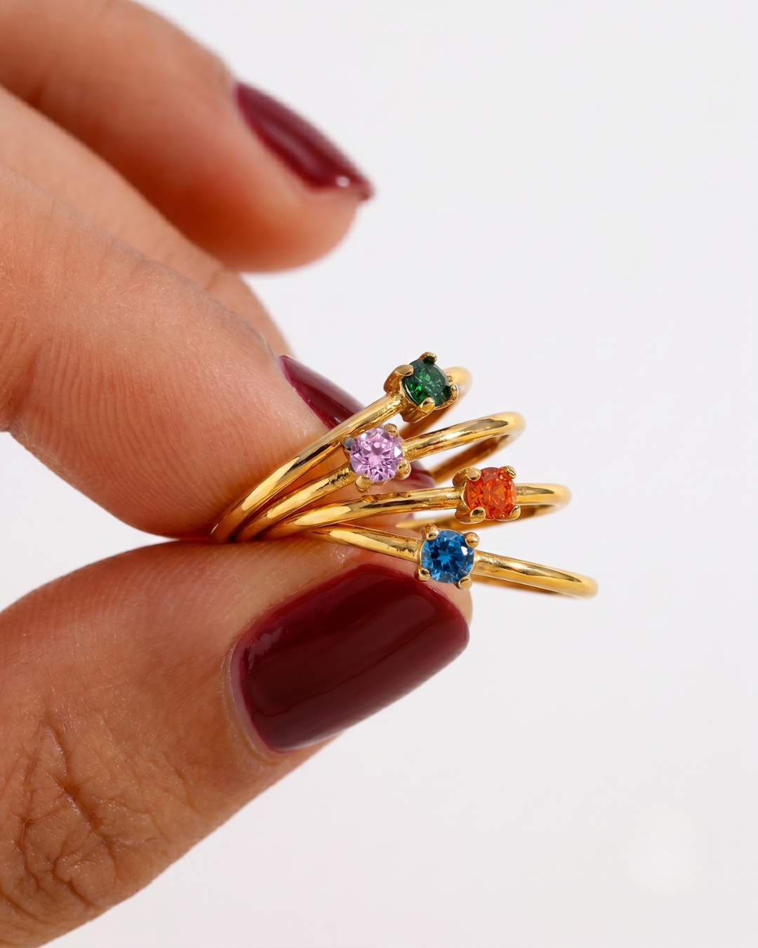 A close-up view of a finger with multiple thin gold stacking rings, each featuring a small, round-cut, multicolored gemstone, including blue, pink, orange, and purple stones.