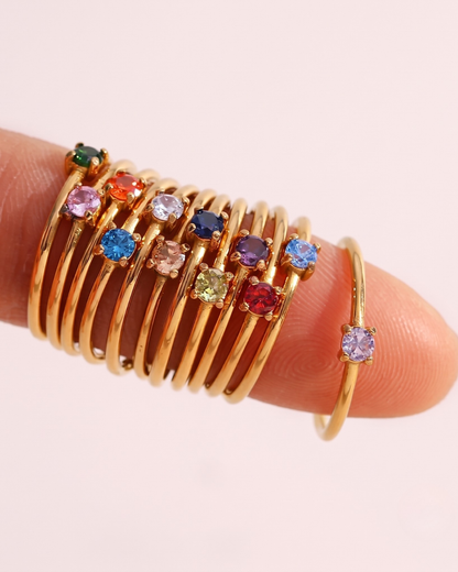 A close-up photograph of a finger wearing a dense stack of thin gold rings, each with a single, small, round-cut gemstone in a variety of vibrant colors.