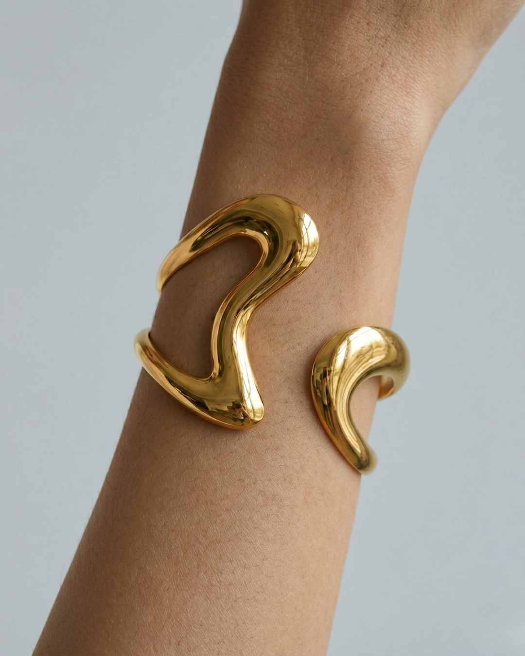 Woman wearing gold open bangle bracelet with fluid line design on wrist, minimal white top, soft natural light — Scilla Bracelet by Clarabelle