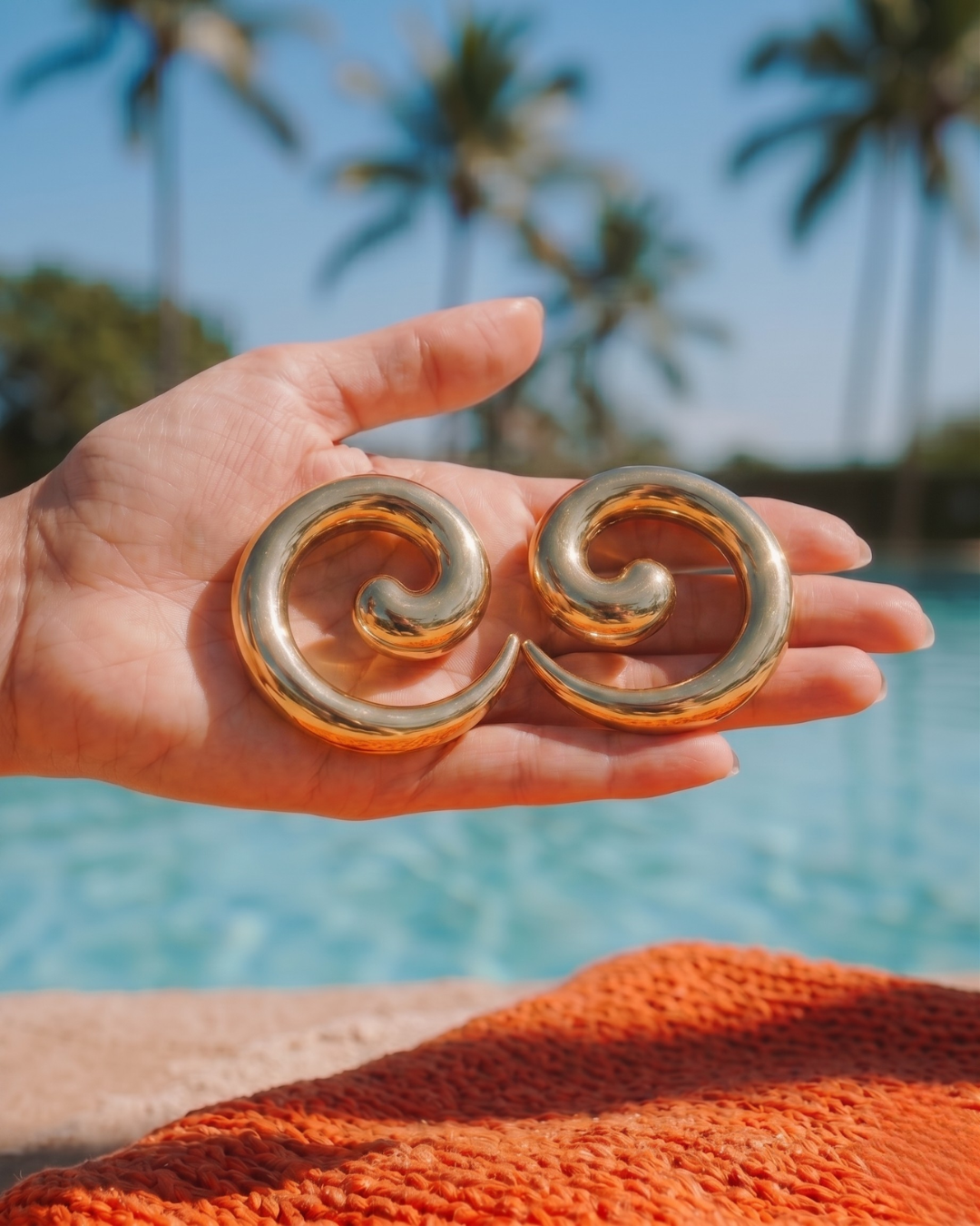 Hand holding gold statement earrings with pool and palm trees in background, summer jewellery look