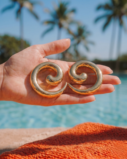 Hand holding gold statement earrings with pool and palm trees in background, summer jewellery look