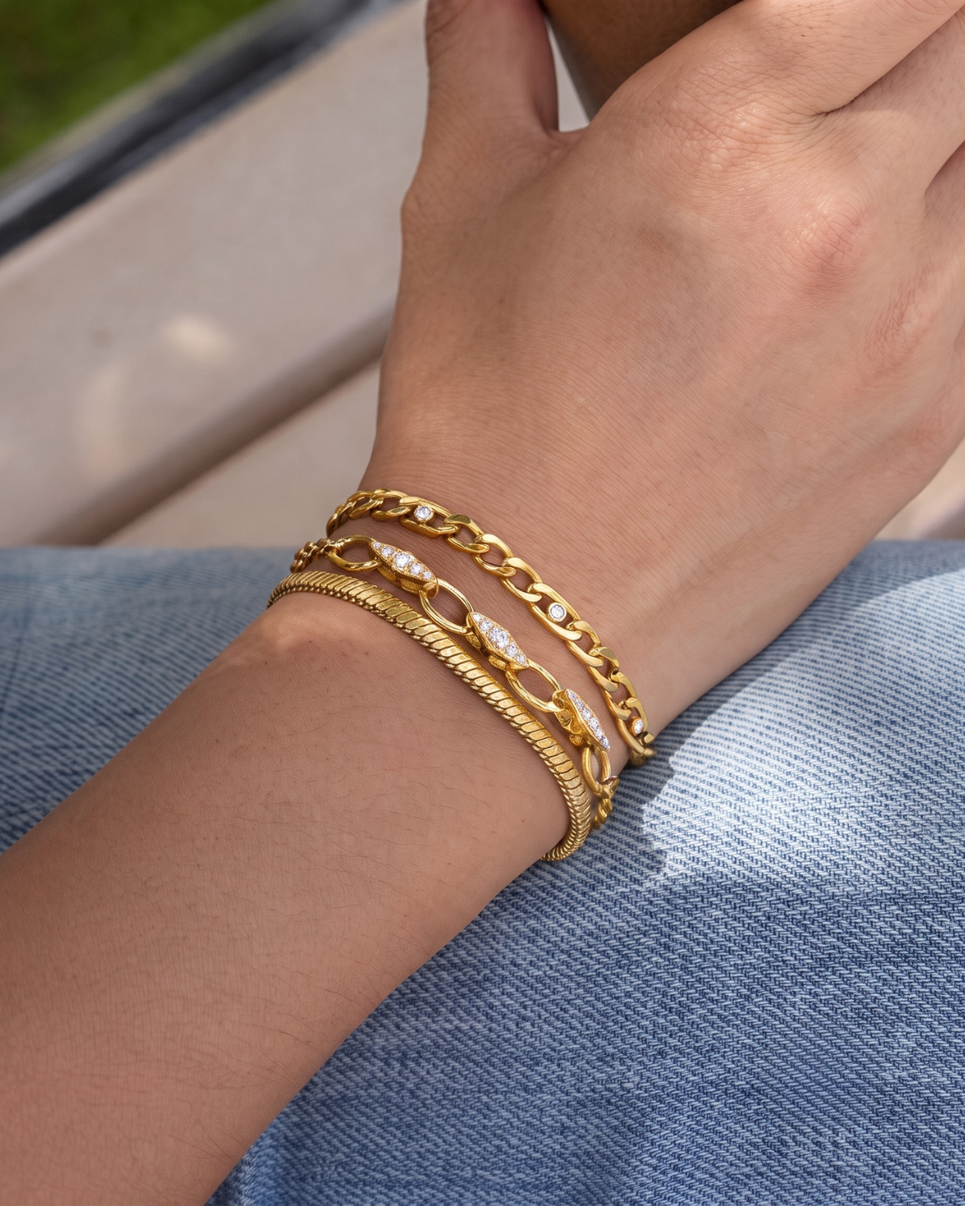 The Wrist Stack worn on the wrist against denim — Marcella Figaro, Chiara pavé rolo, and Lucia snake chain layered in gold finish by Clarabelle