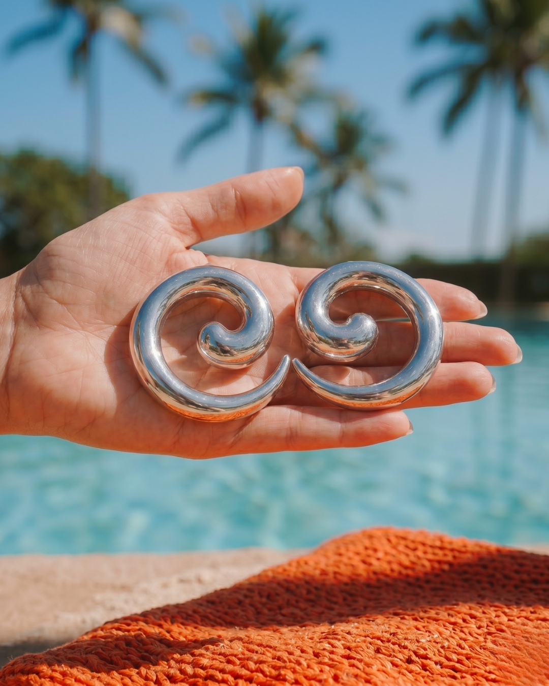 Hand holding silver statement earrings with pool and palm trees in background, summer jewellery look